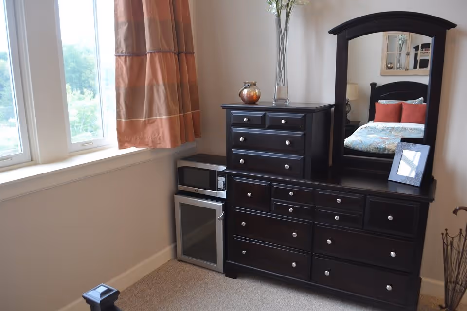 A corner of a bedroom featuring a dark wooden dresser with multiple drawers and a large mirror. On top of the dresser are decorative items including a vase with tall branches and a small framed photo. Next to the dresser is a small microwave and a mini refrigerator. A window with orange and beige curtains lets in natural light.