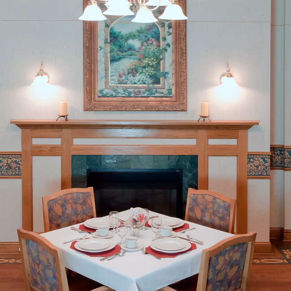 A dining table set for four with white plates, cups, and silverware on red napkins, placed in front of a wooden fireplace with a green tile surround. Above the fireplace is a framed painting of a garden path, flanked by two wall sconces with lit lights and two candles on the mantel.