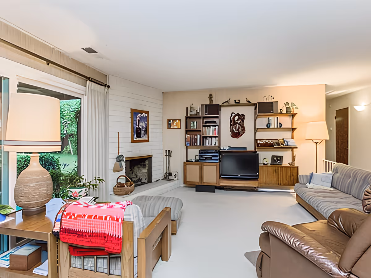 A cozy living room with a large window showing a green garden outside. The room features a wooden bookshelf with books and decorative items, a flat-screen TV, a fireplace, a beige sofa, a brown leather armchair, a wooden side table with a large lamp, and a chair with a red and pink blanket draped over it.