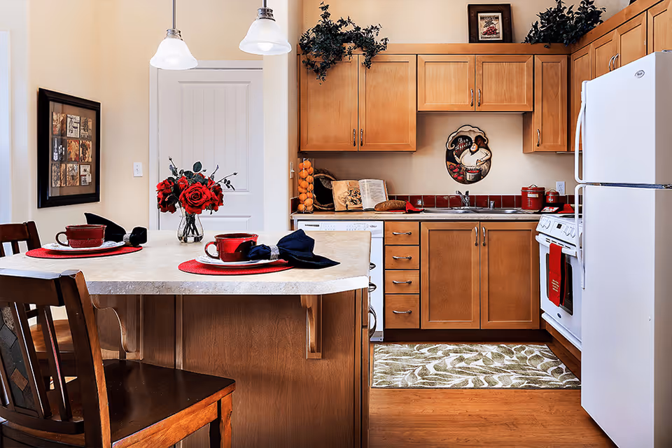 A warm kitchen with a center island set with red place settings and a vase of red flowers, wooden cabinets, white appliances, and decorative red accents.