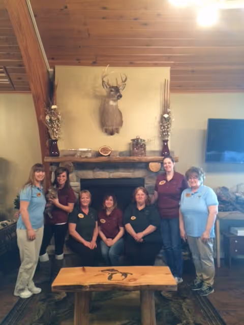 Seven staff members pose in a rustic common room in front of a stone fireplace topped by a mounted deer head.