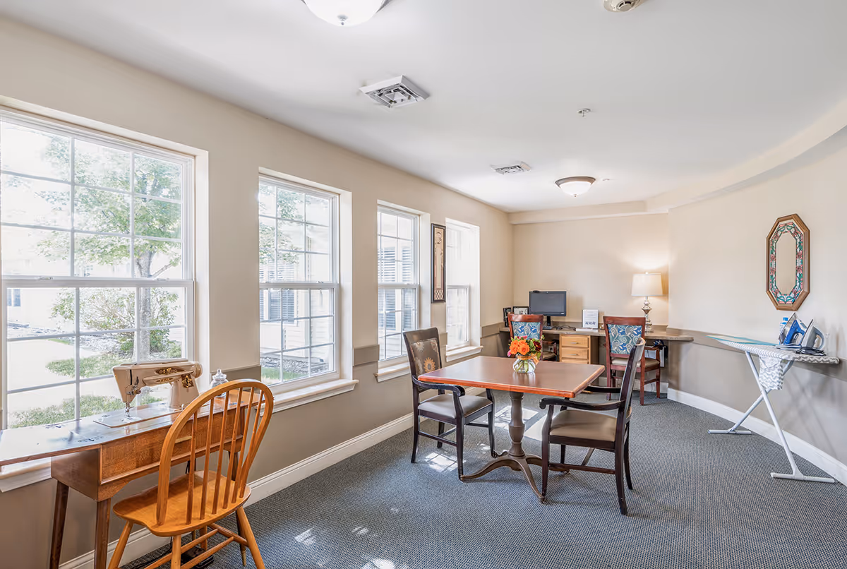 A bright room with three large windows letting in natural light. The room contains a wooden table with four chairs, a small desk with a computer and lamp, an ironing board with an iron, and a vintage sewing machine on a small table by the window. The walls are beige and the carpet is gray.