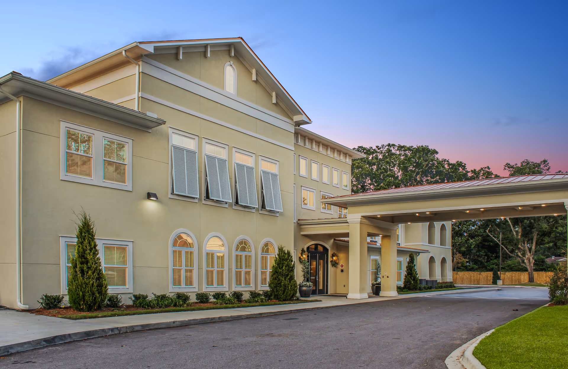 Two-story beige senior living building with a covered entrance and landscaped driveway at dusk.