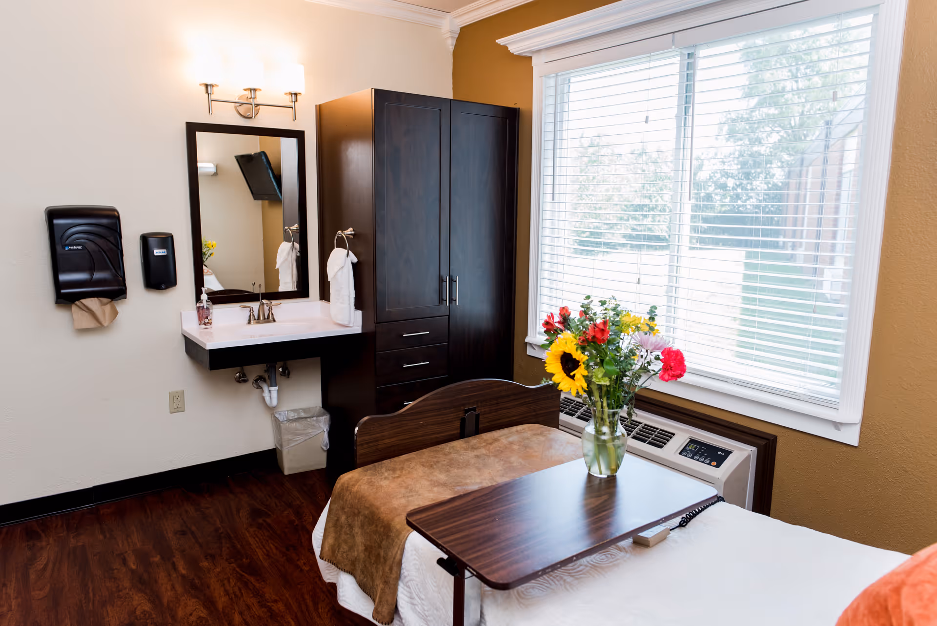 A well-lit room with a single bed covered in white and brown bedding, a wooden overbed table with a vase of colorful flowers, a large window with white blinds, a dark wooden wardrobe, and a wall-mounted sink with a mirror and light fixture above it.