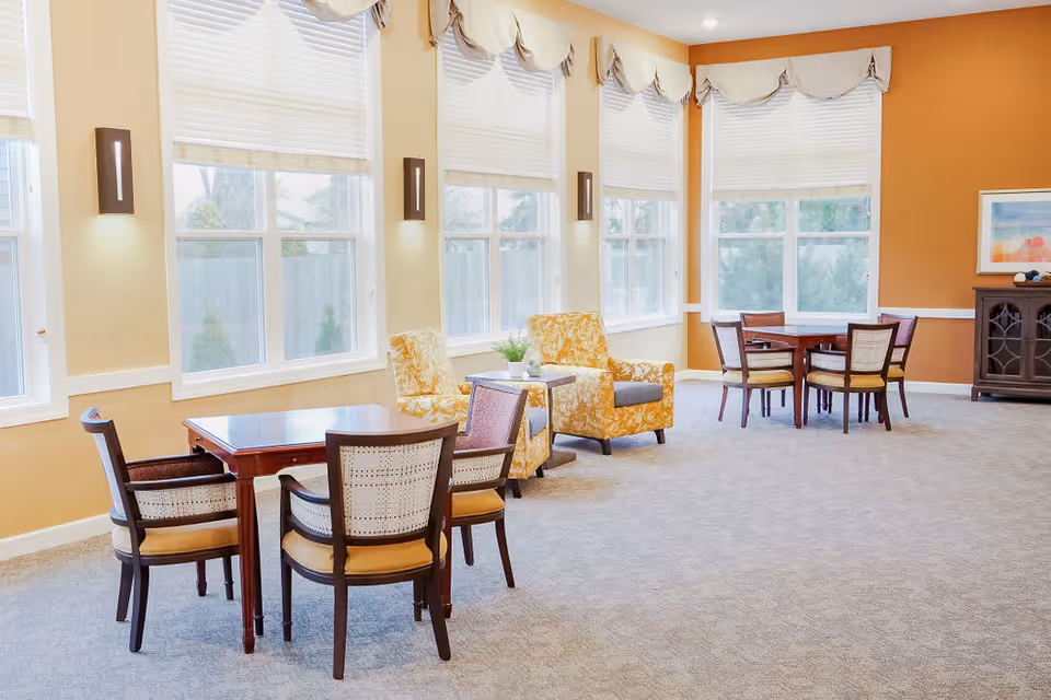 A bright and spacious common area in a senior living facility with large windows covered by white blinds and valances. The room features two sets of wooden tables with four chairs each, and two yellow patterned armchairs with a small table between them. The walls are painted in warm tones of beige and orange, and there is a wooden cabinet with a framed artwork above it in the corner.