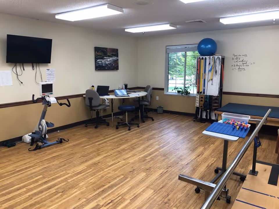 A bright interior rehab/exercise room with a stationary bike, parallel bars, therapy table, exercise equipment and a table with chairs by a window.