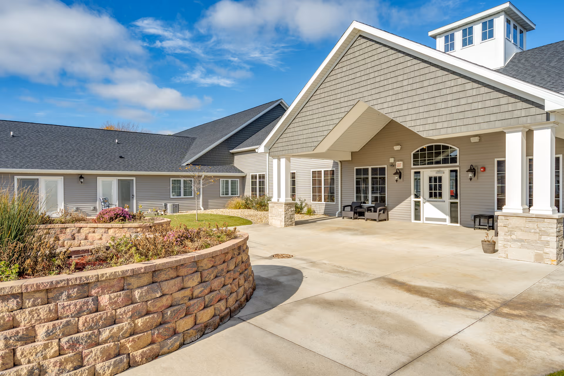 Exterior view of Villas of Holly Brook Assisted Living & Memory Care facility in Pekin, IL, showing the entrance with a covered porch, stone pillars, and a landscaped area with a curved stone retaining wall under a partly cloudy blue sky.