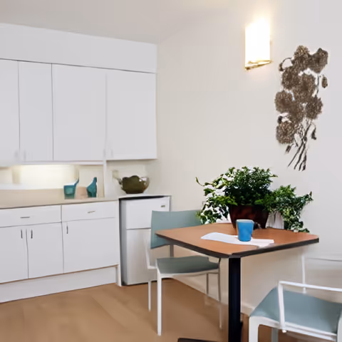A small dining area with a wooden table and two chairs with light blue cushions. On the table, there is a blue cup and a white napkin. Behind the table is a white kitchenette with cabinets, a small refrigerator, and decorative items. A potted green plant is placed on the table, and a wall-mounted light fixture and a decorative wall art piece are visible on the white wall.