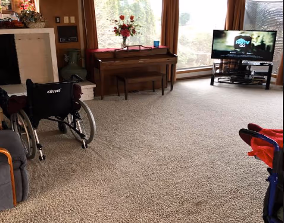 A living room with beige carpet, a wheelchair, a fireplace, a wooden piano with a vase of flowers on top, large windows with brown curtains, and a television on a stand showing HGTV GO.