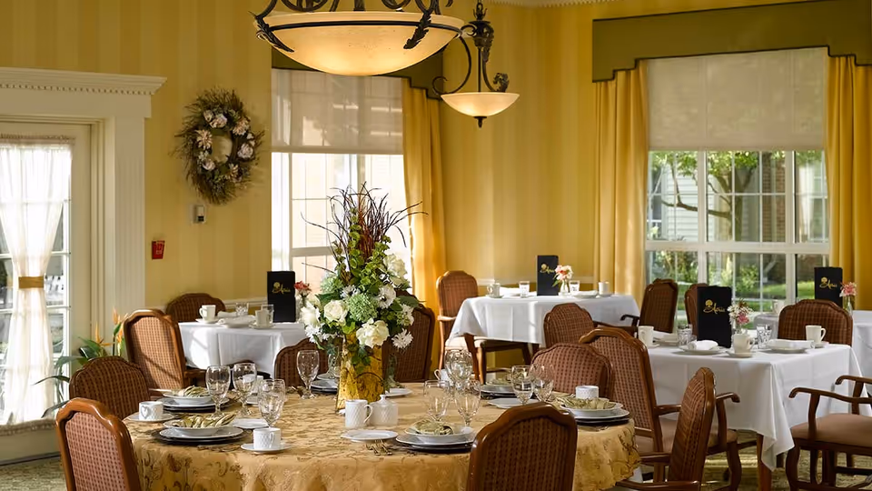 A bright dining room with several round and rectangular tables covered with white and gold tablecloths. Each table is set with plates, cups, glasses, and silverware. A large floral centerpiece is on the round table in the foreground. The room has large windows with yellow curtains, and a door with sheer white curtains. Warm lighting fixtures hang from the ceiling.
