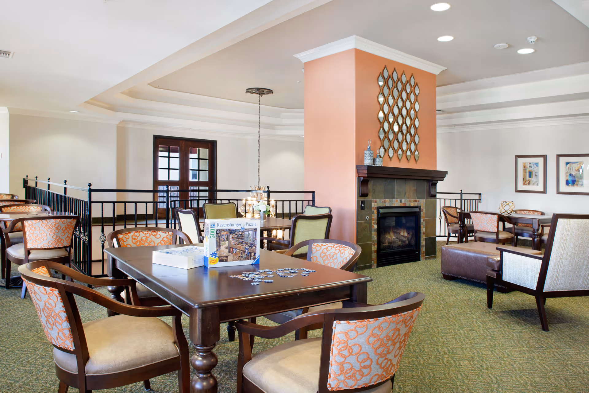 A spacious common area in a senior living facility featuring multiple tables and chairs with patterned upholstery. A dark wooden table in the foreground has a partially completed Ravensburger puzzle on it. The room includes a fireplace with a decorative mirror above it, framed artwork on the walls, and a green carpeted floor. The ceiling has recessed lighting and a chandelier hangs over one of the tables.