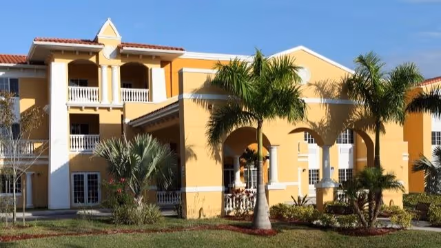 Exterior view of a yellow two-story assisted living community building with white trim, arches, balconies, and palm trees in the landscaped front yard under a clear blue sky.