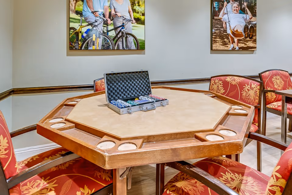 A wooden octagonal game table with cup holders and a poker chip set in an open case on top. Surrounding the table are red cushioned chairs with floral patterns. Two framed photos hang on the wall behind the table, one showing two people with bicycles and the other showing an elderly person on a swing.