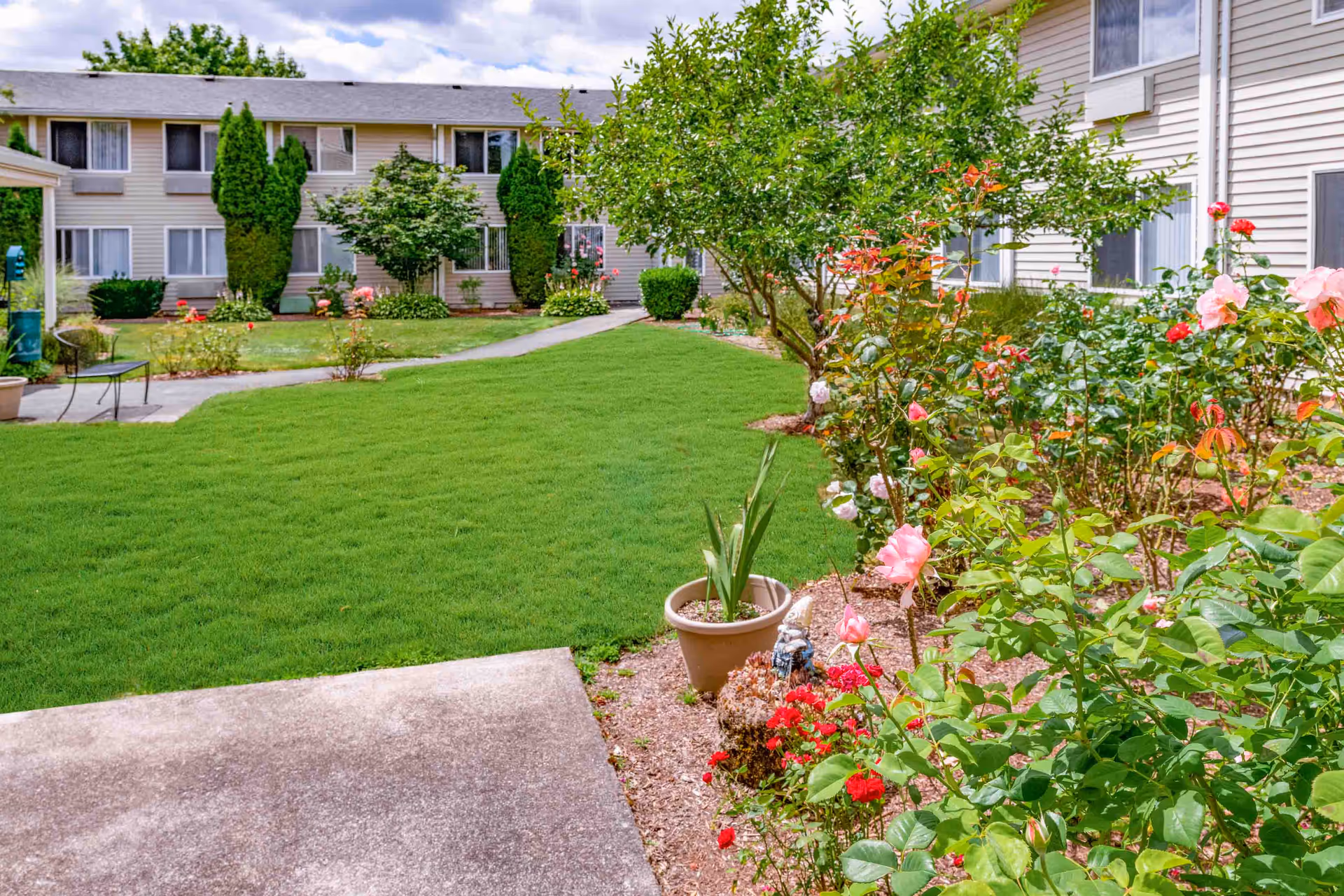Outdoor garden area at Holiday Edgewood Downs featuring a well-maintained lawn, blooming rose bushes, a potted plant, a small garden gnome, and a paved walkway leading to a two-story residential building with beige siding and multiple windows.