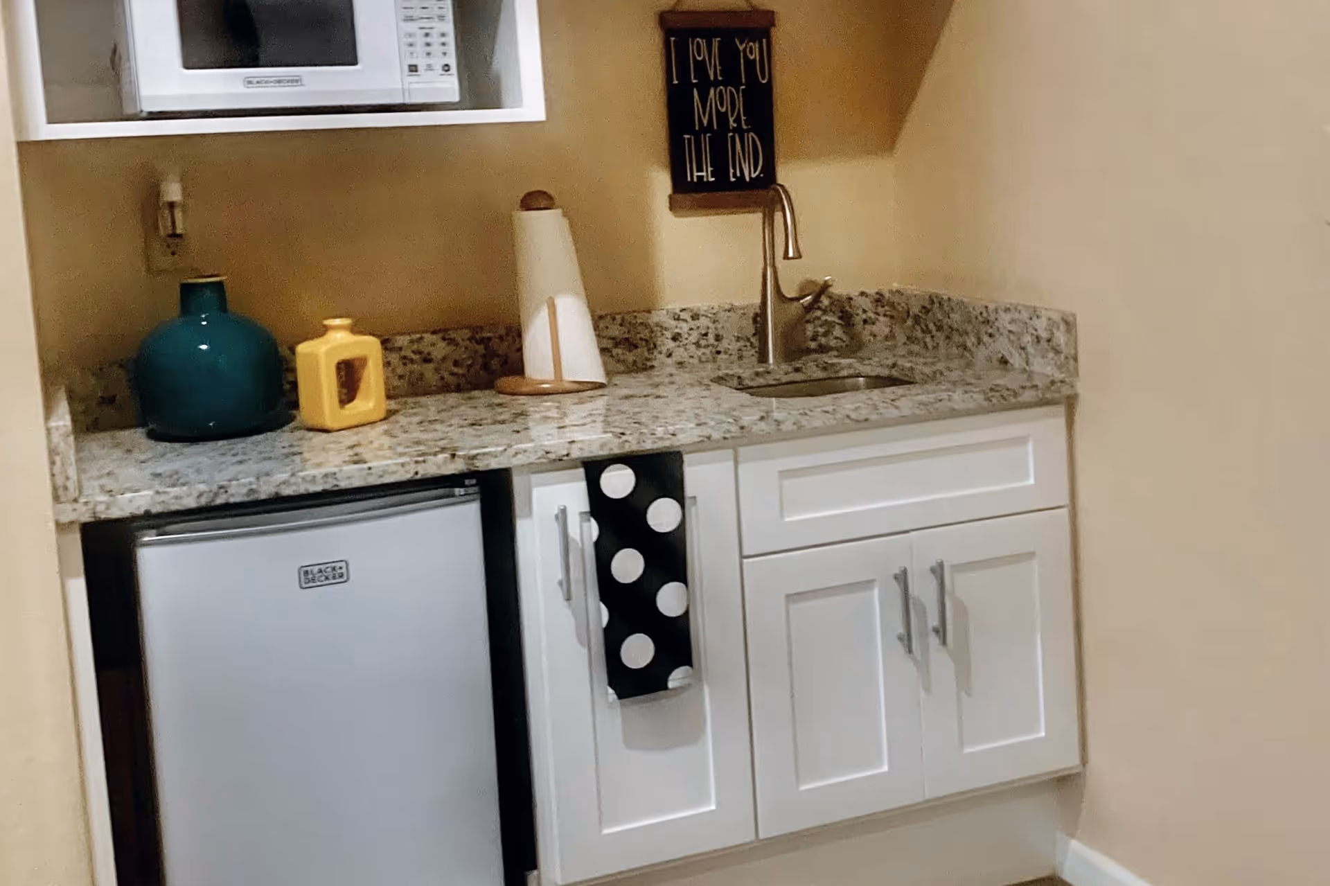 A small kitchenette area with a granite countertop, a stainless steel sink with a faucet, a mini refrigerator, a microwave, and white cabinets. There are decorative items on the counter including a teal vase, a yellow container, and a paper towel holder. A black towel with white polka dots hangs from one of the cabinet handles. A small wall sign above the sink reads 'I LOVE YOU MORE THE END.'