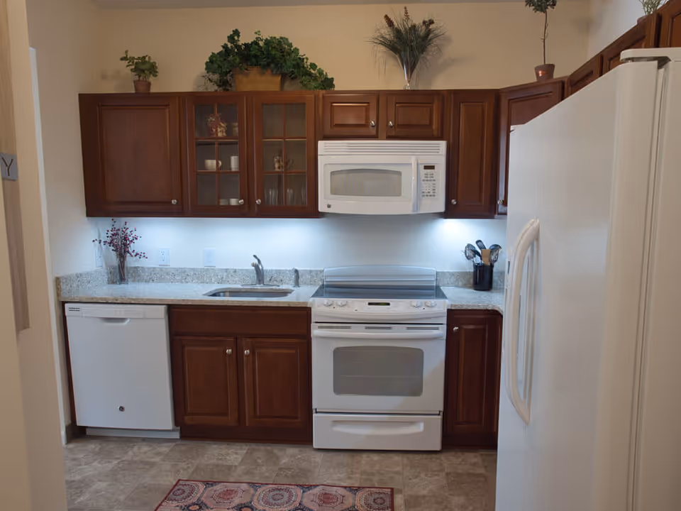A kitchen with wooden cabinets, a white dishwasher, a white stove with an oven, a white microwave above the stove, a white refrigerator, a granite countertop with a sink, and decorative plants on top of the cabinets.