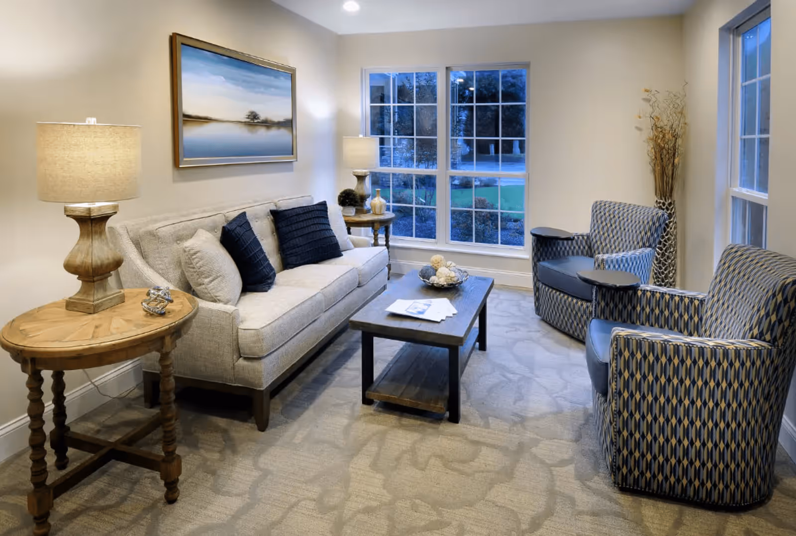 A cozy living room with a beige sofa adorned with dark and light cushions, two patterned armchairs, a wooden coffee table with decorative items and papers, two wooden side tables each with a lamp, a large window showing an outdoor view, and a framed landscape painting on the wall.