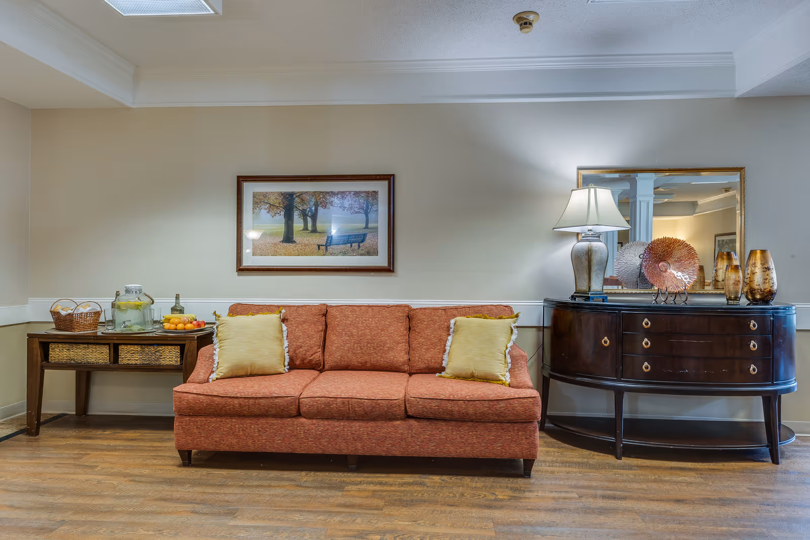 A cozy seating area featuring a rust-colored sofa with two yellow pillows, a wooden side table with a basket, a glass jar of lemon water, and a bowl of fruit. On the right, there is a dark wooden console table with a lamp, decorative vases, and a large mirror above it. A framed picture of a park bench under trees hangs on the wall above the sofa. The room has wooden flooring and beige walls.
