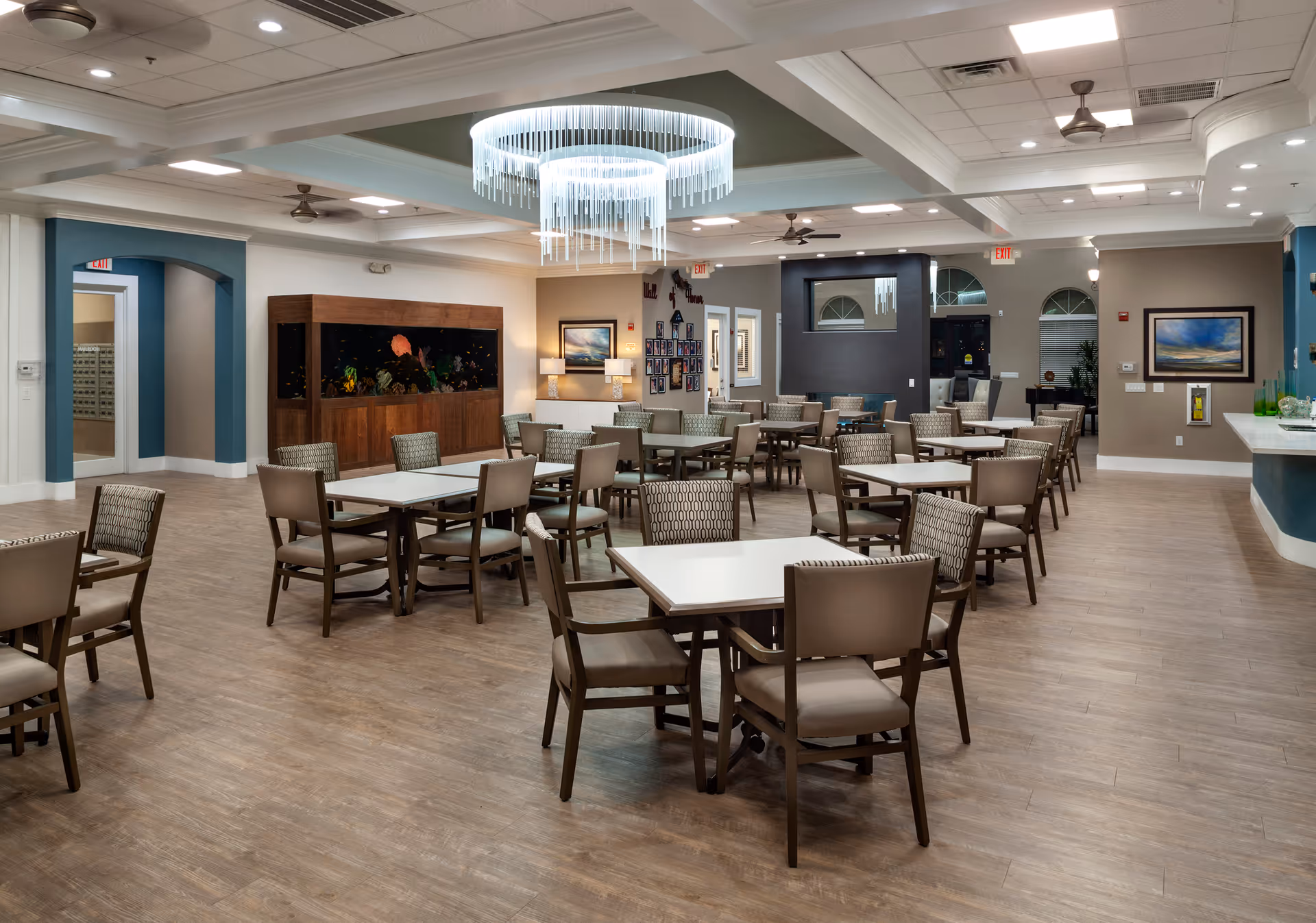 Spacious dining and activity room with multiple tables and chairs, a large aquarium and a modern chandelier.