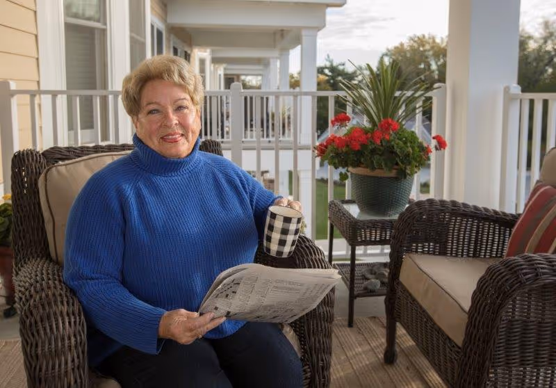 An elderly woman wearing a blue sweater sits on a wicker chair on a porch, holding a newspaper and a black-and-white checkered mug. There is a small table with a potted plant with red flowers next to her, and another wicker chair with striped cushions is visible. The porch has white railings and overlooks a green outdoor area.