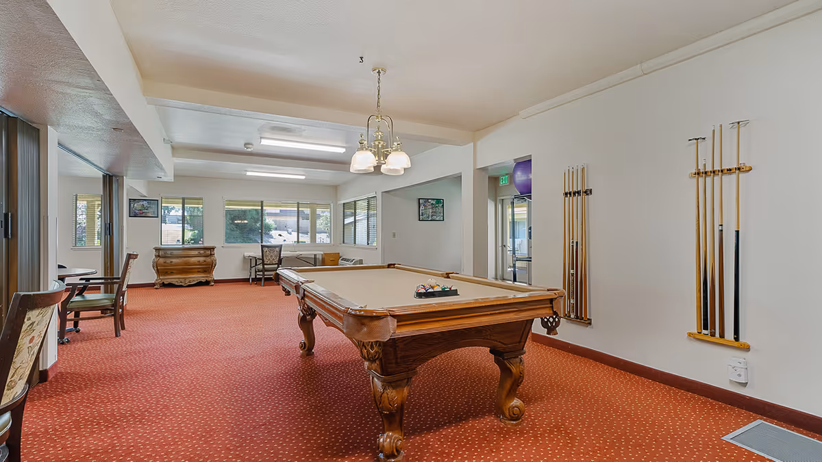 A spacious game room with a pool table in the center, pool cues mounted on the wall, several chairs and tables along the sides, large windows letting in natural light, and a red carpeted floor.