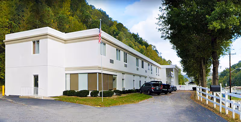Exterior view of a two-story white building with several windows, an American flag on a flagpole in front, a few parked cars, and a white fence along the driveway. The building is surrounded by trees and hills under a partly cloudy sky.