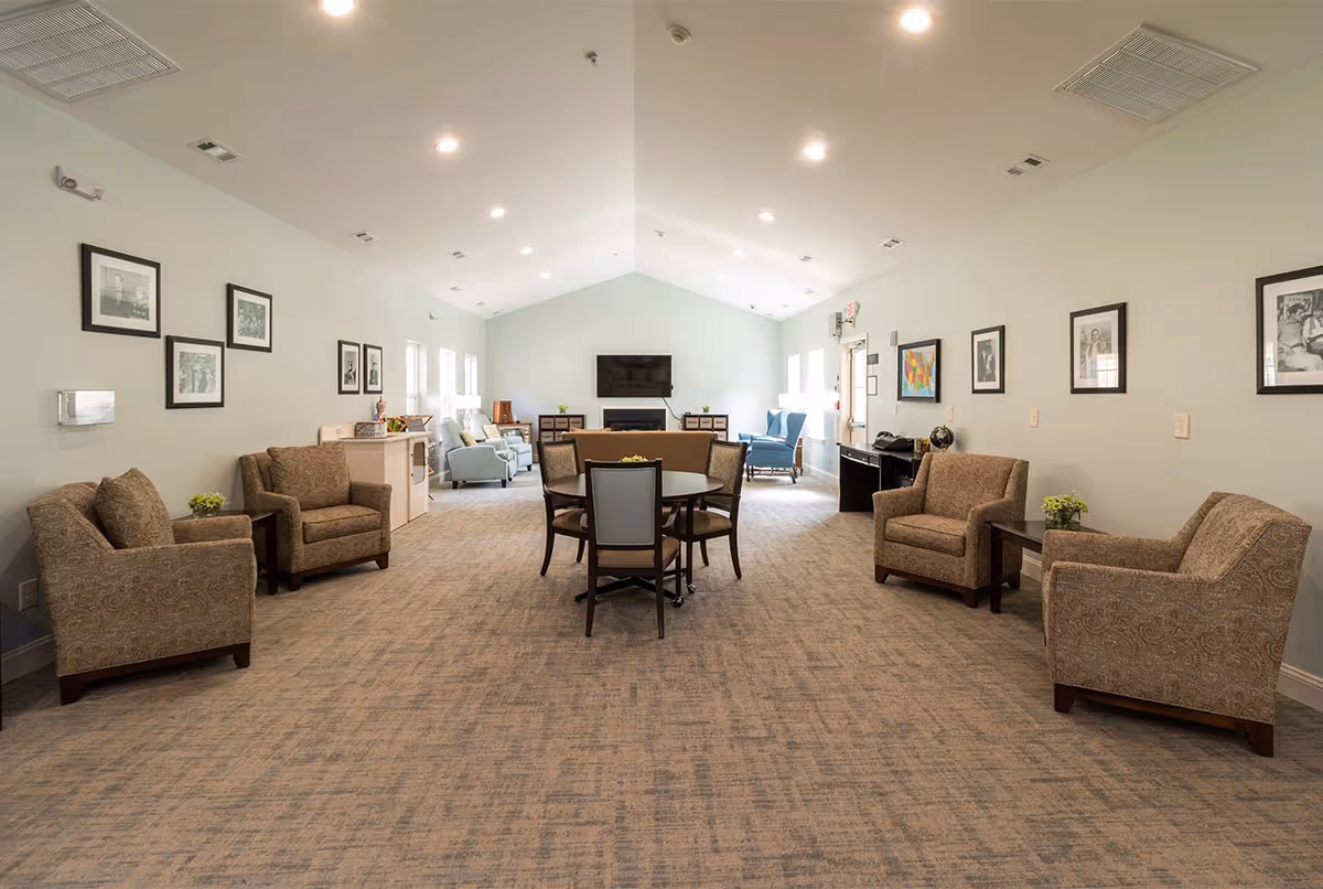 A spacious senior living common area with beige patterned armchairs arranged along the walls, a round table with four chairs in the center, and a TV mounted on the far wall above a fireplace. The room has light-colored walls adorned with framed black and white photographs, several windows letting in natural light, and recessed ceiling lights.