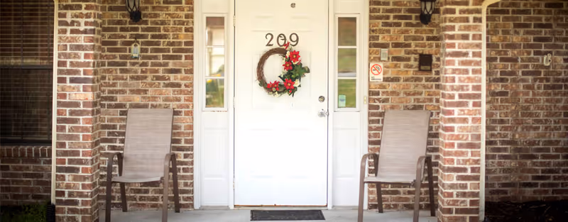 Front entrance of a brick building with a white door numbered 209, decorated with a wreath featuring red flowers. Two beige chairs are placed on either side of the door on the porch.