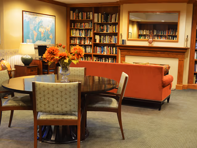 Cozy common room with a round table and chairs, a vase of orange flowers, an orange sofa, bookshelves and a fireplace.