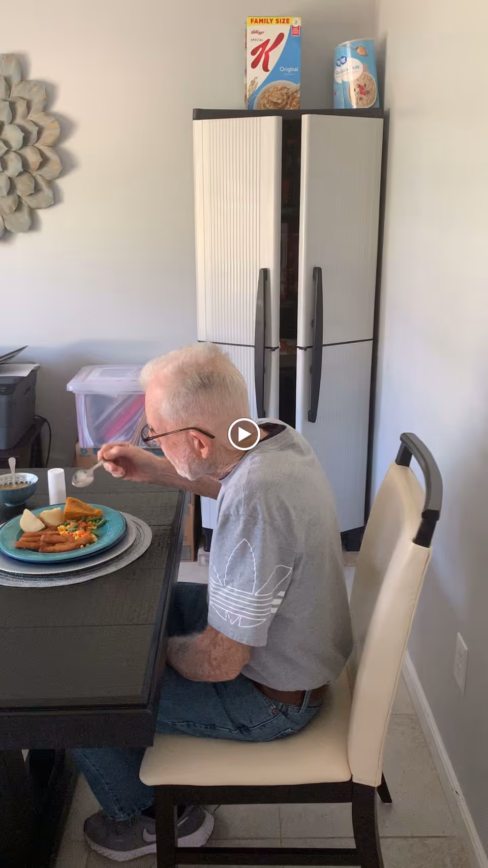 An elderly man with white hair and glasses is sitting at a dining table eating a meal. The table has a plate with various foods including vegetables and what appears to be fried items. Behind him is a white cabinet with two cereal boxes on top. The room has light-colored walls and a decorative wall piece is partially visible on the left.