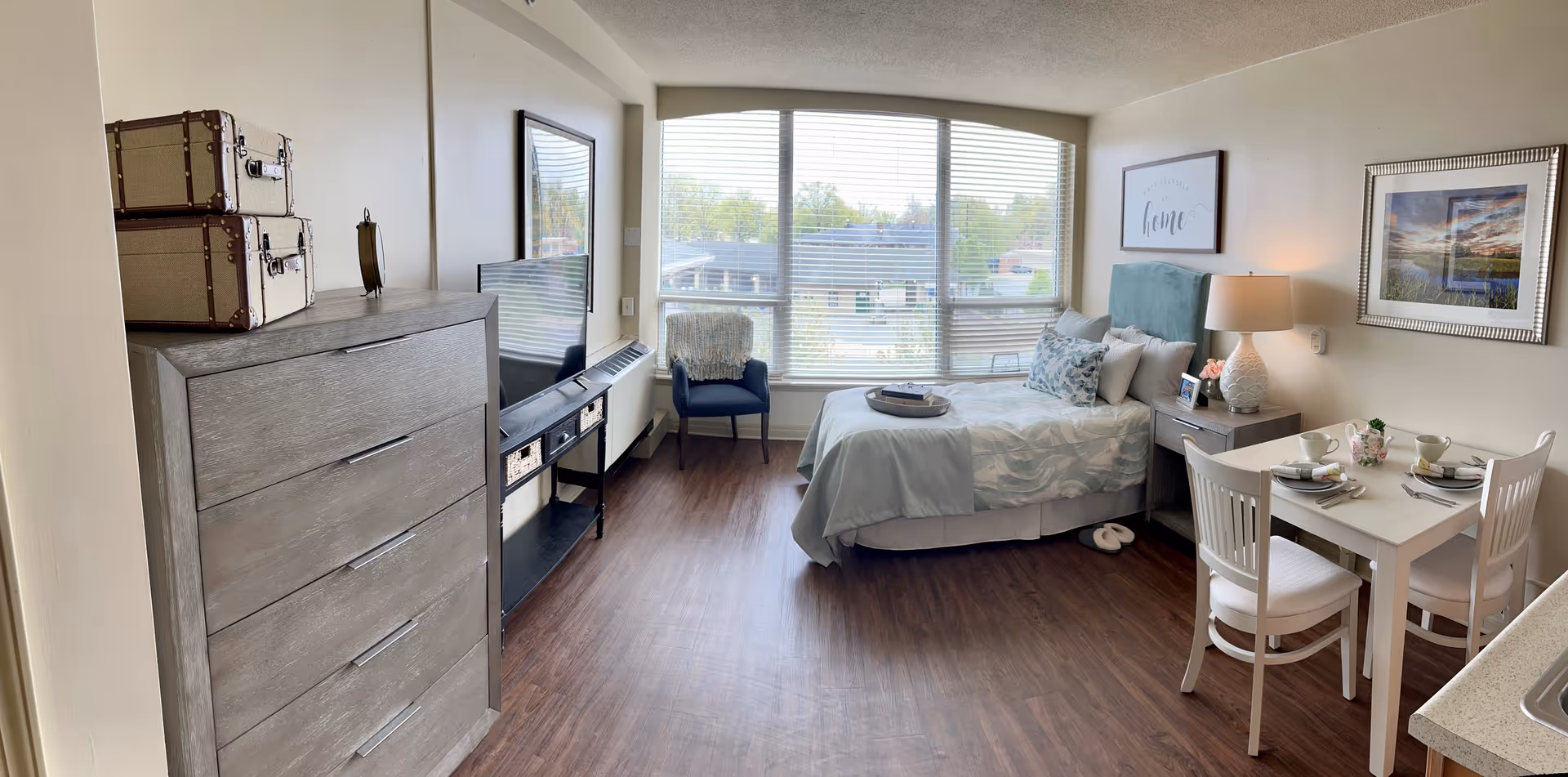 A bright and cozy senior living studio apartment featuring a bed with light blue bedding and decorative pillows, a nightstand with a lamp and framed photo, a small dining table set for two, a dresser with stacked suitcases on top, a flat-screen TV on a stand, and a window with blinds letting in natural light. The room has wood flooring and neutral-colored walls with framed artwork.