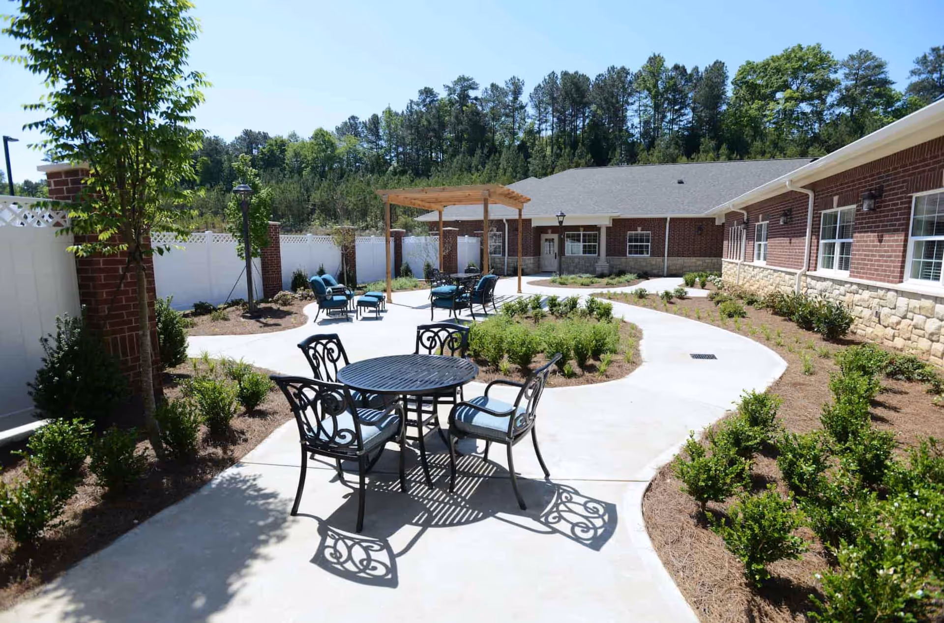 Outdoor patio area at the Reserve at Towne Lake featuring a curved concrete walkway, metal table with four chairs, additional seating under a wooden pergola, surrounded by landscaped bushes and trees, with a brick and stone building in the background.