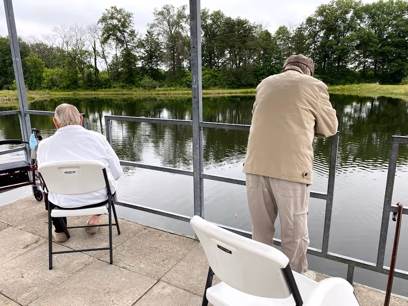 Two elderly men at a lakeside fishing spot. One man is sitting on a white folding chair with a walker beside him, while the other man is standing and leaning on a metal railing, looking out over the water. The background shows a calm lake with trees along the far shore under a cloudy sky.