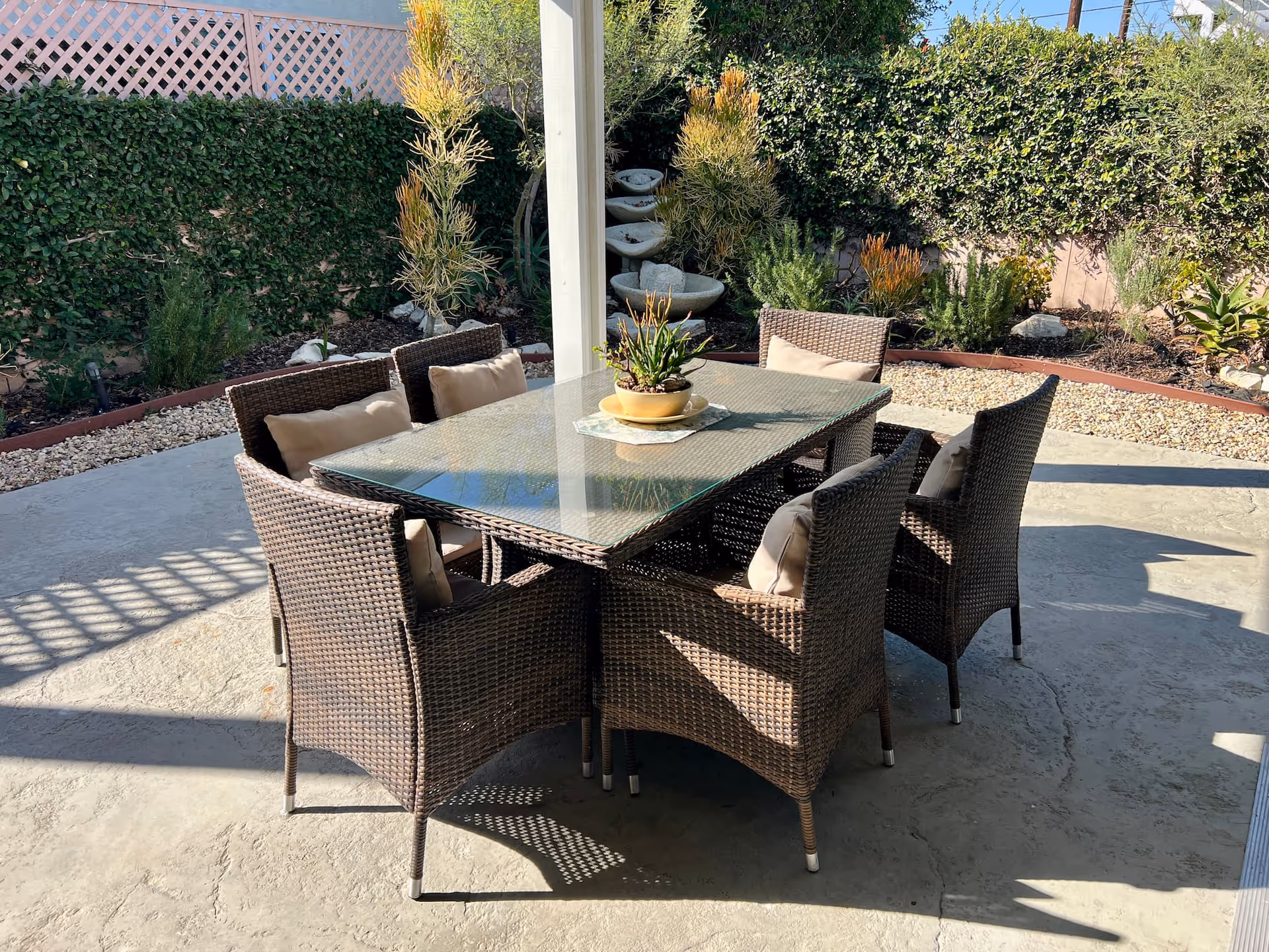 Outdoor patio area with a glass-top table surrounded by six wicker chairs with beige cushions. The patio is shaded by a pergola, and there are various plants and shrubs along the perimeter, including a tiered stone water feature in the background.