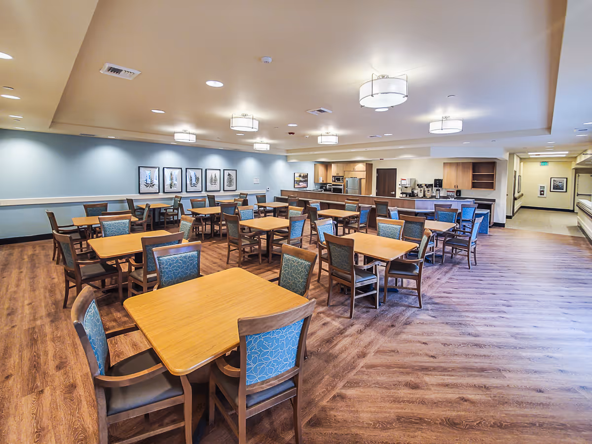 A spacious dining room with multiple wooden tables and chairs with blue patterned upholstery. The room has wood flooring, soft blue walls with framed artwork, and ceiling lights. In the background, there is a kitchen area with appliances and a coffee station.
