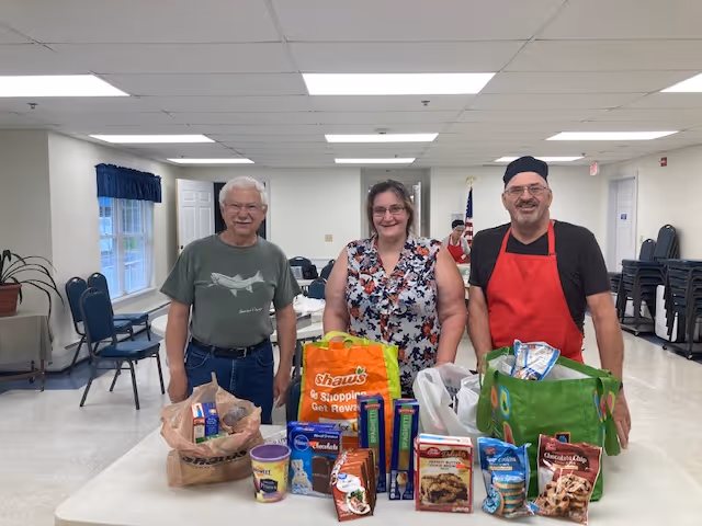 Three adults standing behind a table filled with grocery bags and various food items in a bright, spacious room with chairs and tables in the background.