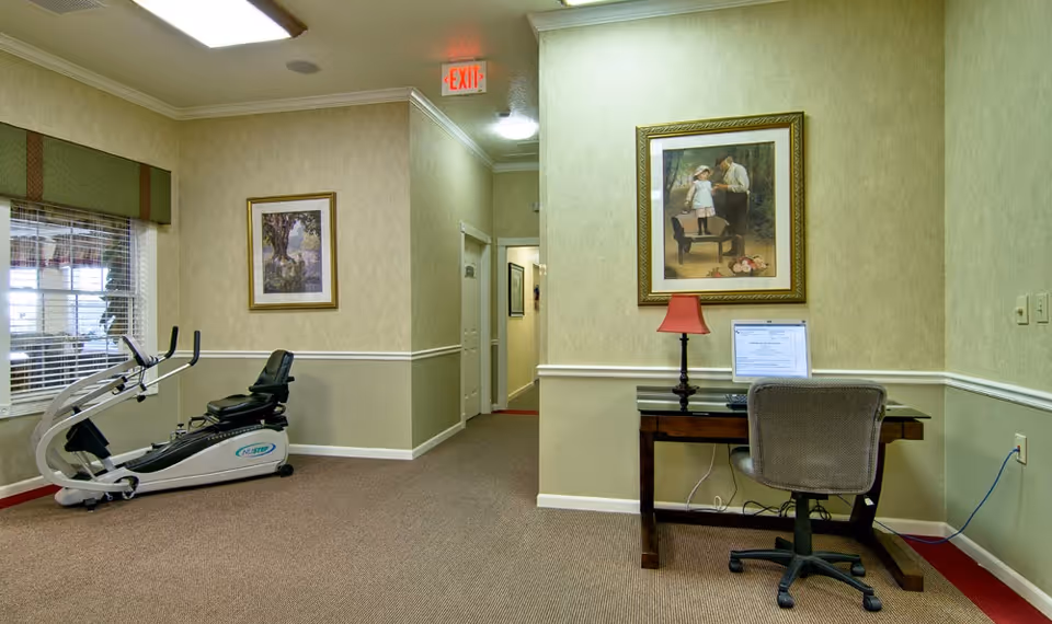 Interior view of a senior living facility room with beige walls and carpeted floor. On the left side, there is a recumbent exercise bike near a window with blinds and a framed picture above it. On the right side, there is a small wooden desk with a computer monitor, a red lamp, and an office chair. Above the desk hangs a framed painting of a man and a child. A hallway with doors and an exit sign is visible in the background.