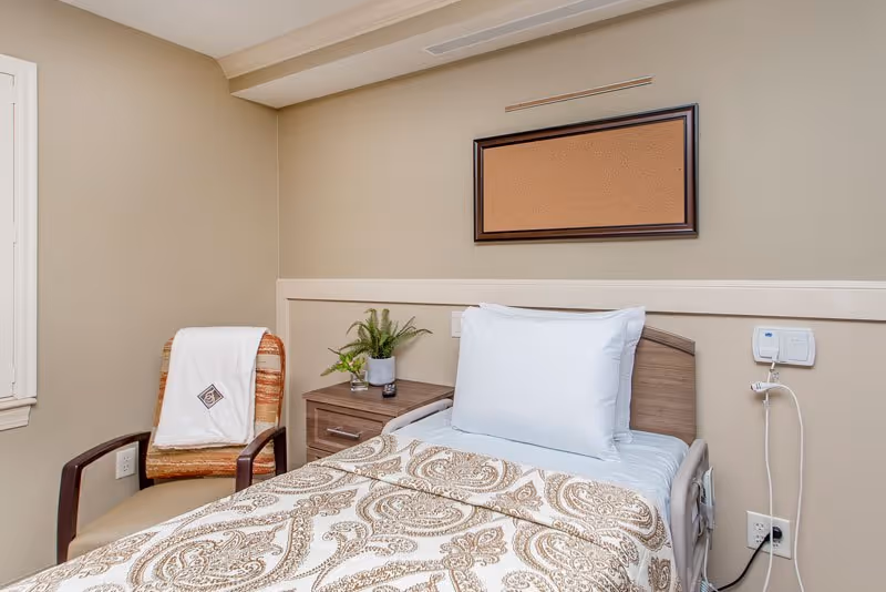 A neatly made single bed with a patterned beige and white blanket and a large white pillow in a small room. Next to the bed is a wooden nightstand with a small potted plant and a glass of water. A cushioned chair with a white blanket draped over the back is positioned near the bed. The walls are beige, and there is a cork bulletin board mounted above the bed. A call button and electrical outlets are visible on the wall beside the bed.