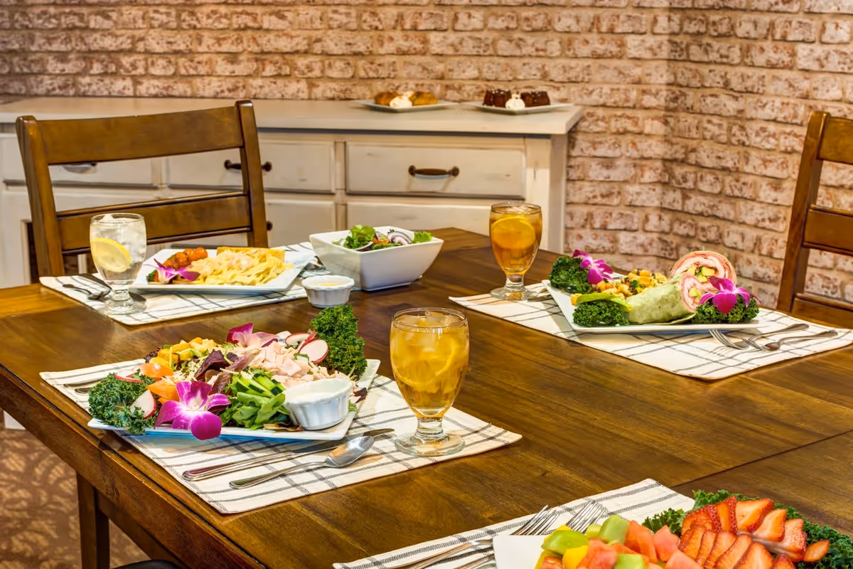 A wooden dining table set with plates of fresh salads, wraps, fruit, and glasses of iced tea and water with lemon. The table is set with checkered placemats and silverware. In the background, there is a white cabinet against a brick wall with plates of desserts on top.