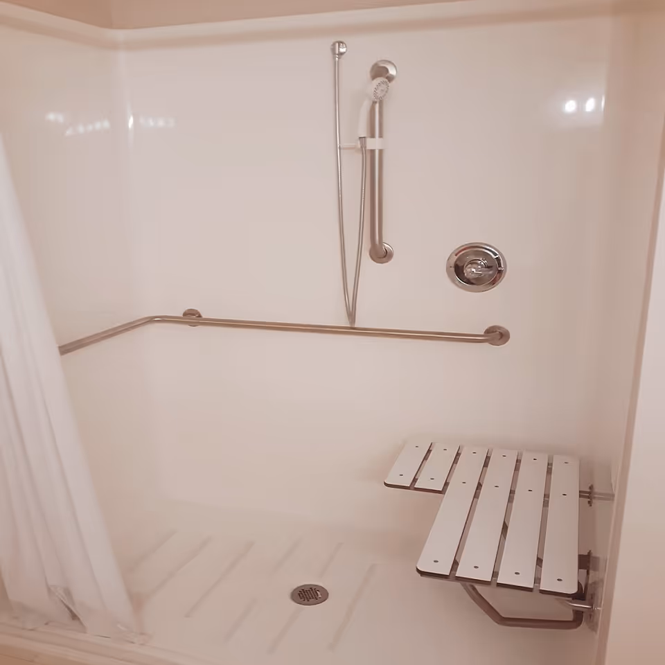 Accessible shower area with a white fold-down bench, a handheld showerhead attached to a vertical grab bar, a horizontal grab bar along the wall, and a white shower curtain partially visible on the left side.
