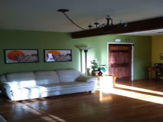 A living room with light green walls, a white sofa, two framed pictures on the wall, a floor lamp, a wooden door, and sunlight streaming through windows onto the wooden floor.