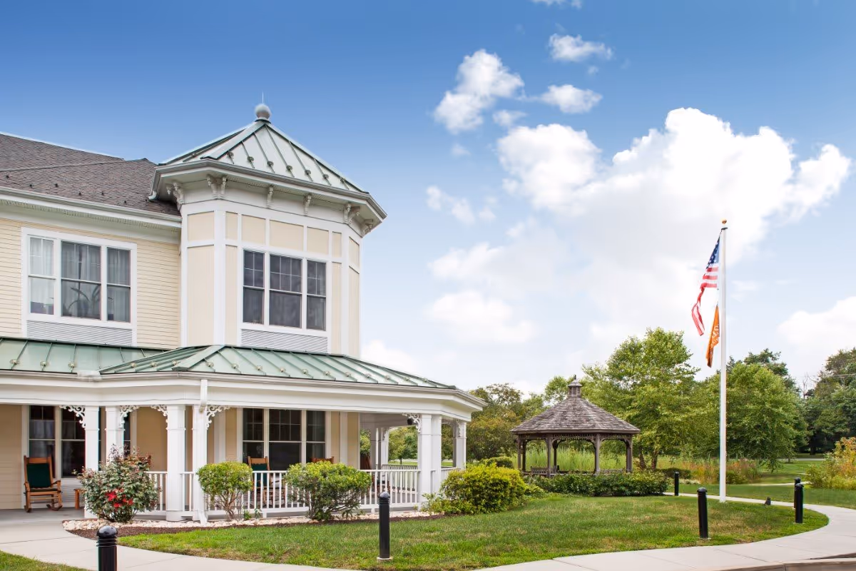 Exterior view of a senior living facility building with a covered porch featuring rocking chairs, surrounded by green bushes and a well-maintained lawn. There is a gazebo in the background and an American flag along with another flag on a flagpole near the sidewalk. The sky is blue with scattered clouds.