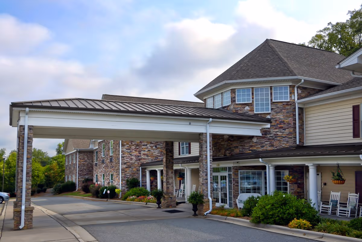 Exterior view of Arbor Ridge At Kernersville senior living facility showing a covered entrance with stone pillars, a driveway, and a building with stone and siding facade, windows, and rocking chairs on the porch.