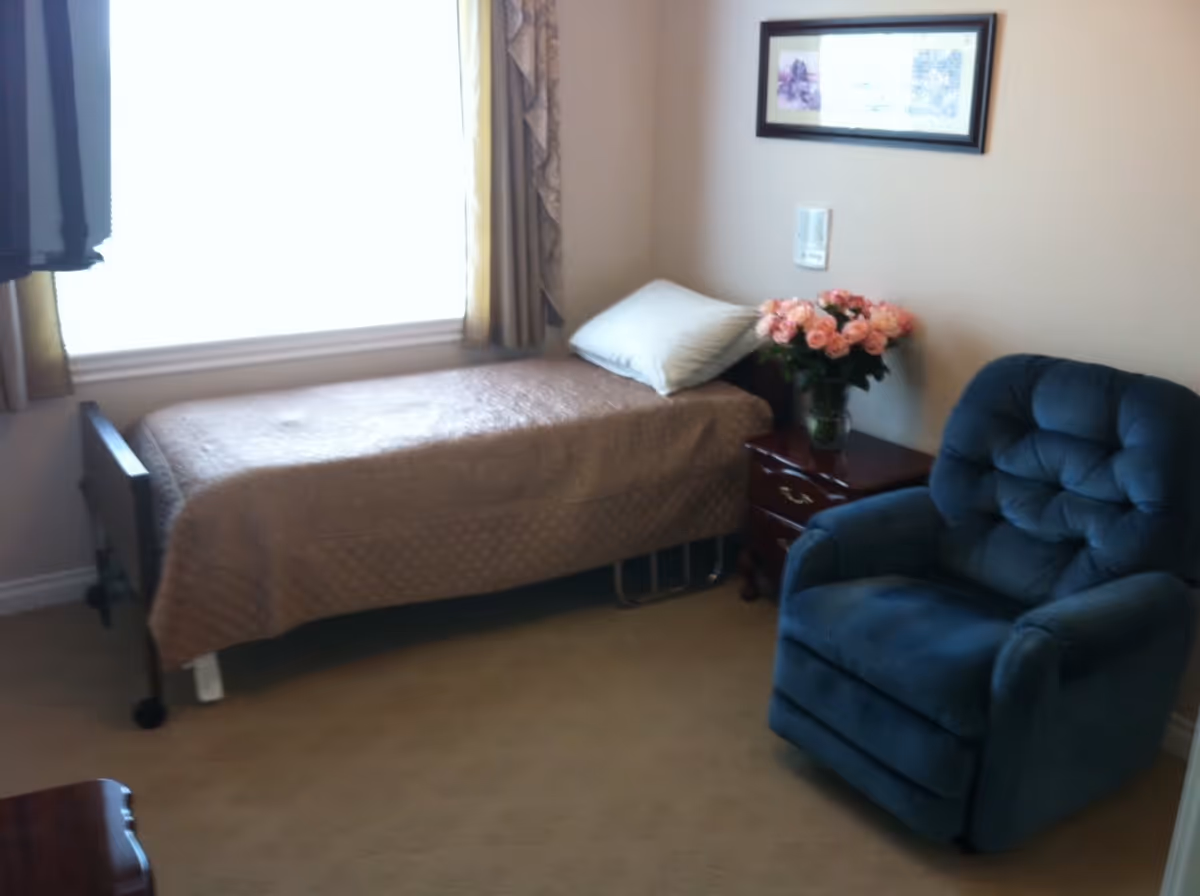 A small bedroom with a single bed covered with a beige quilt, a white pillow, a wooden nightstand with a vase of pink flowers, a blue upholstered armchair, and a framed picture on the wall. A window with curtains is behind the bed.