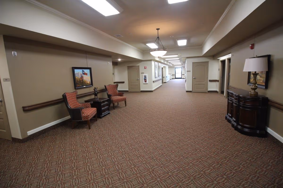 A wide hallway in a senior living facility with patterned carpet and beige walls. The hallway features two upholstered chairs with a small table between them, a framed painting on the wall, a dark wooden sideboard with a lamp, and multiple doors along the corridor. Ceiling lights and a hanging light fixture illuminate the space.