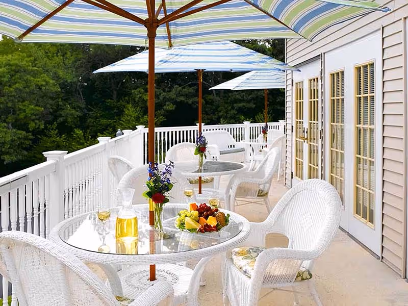 Outdoor balcony patio with white wicker tables and chairs under striped umbrellas, tables set with fruit and drinks.