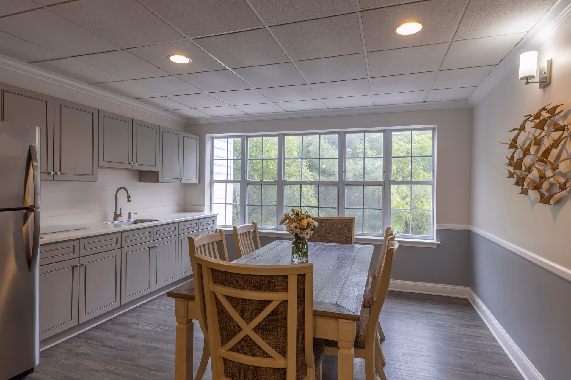 Bright dining area with a wooden table and chairs, gray kitchen cabinets and a large window overlooking greenery.