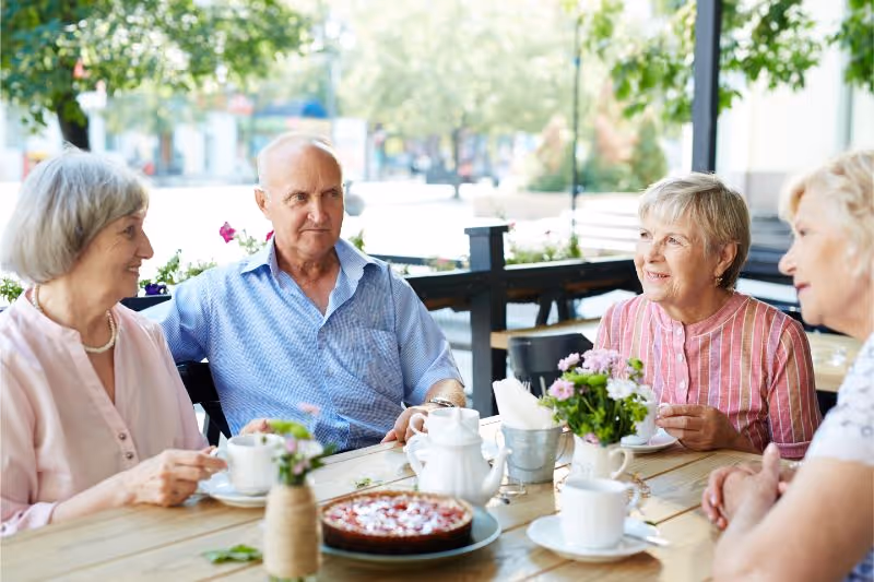 Four elderly people sitting around a wooden table outdoors, enjoying tea and cake, with flowers on the table and greenery in the background.