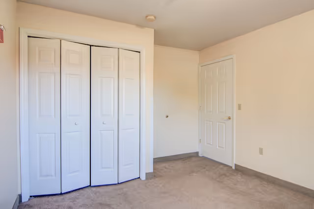 Empty room with beige carpet, cream-colored walls, a white door on the right, and a white bi-fold closet door on the left.