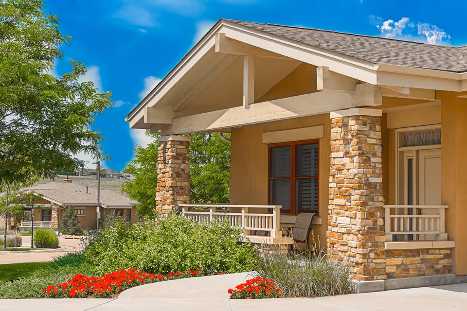 Exterior view of a senior living facility building with stone pillars and a covered porch. There are green bushes and red flowers along the sidewalk leading to the entrance. Another similar building is visible in the background under a bright blue sky with some clouds.